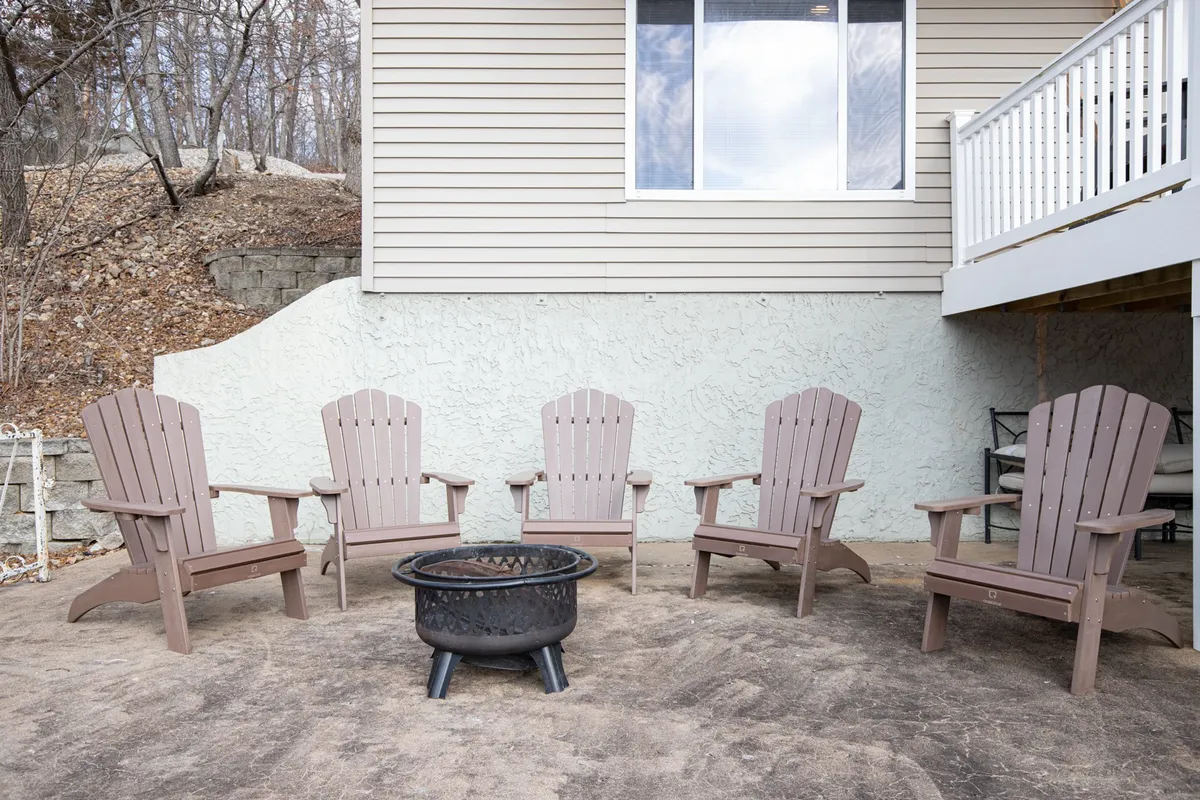 A cozy patio scene with Adirondack chairs encircling a firepit. The fire is lit, creating a warm glow ideal for storytelling, s’mores, and evening relaxation. The exterior of the house is visible in the background, reinforcing the connection to the indoor space.