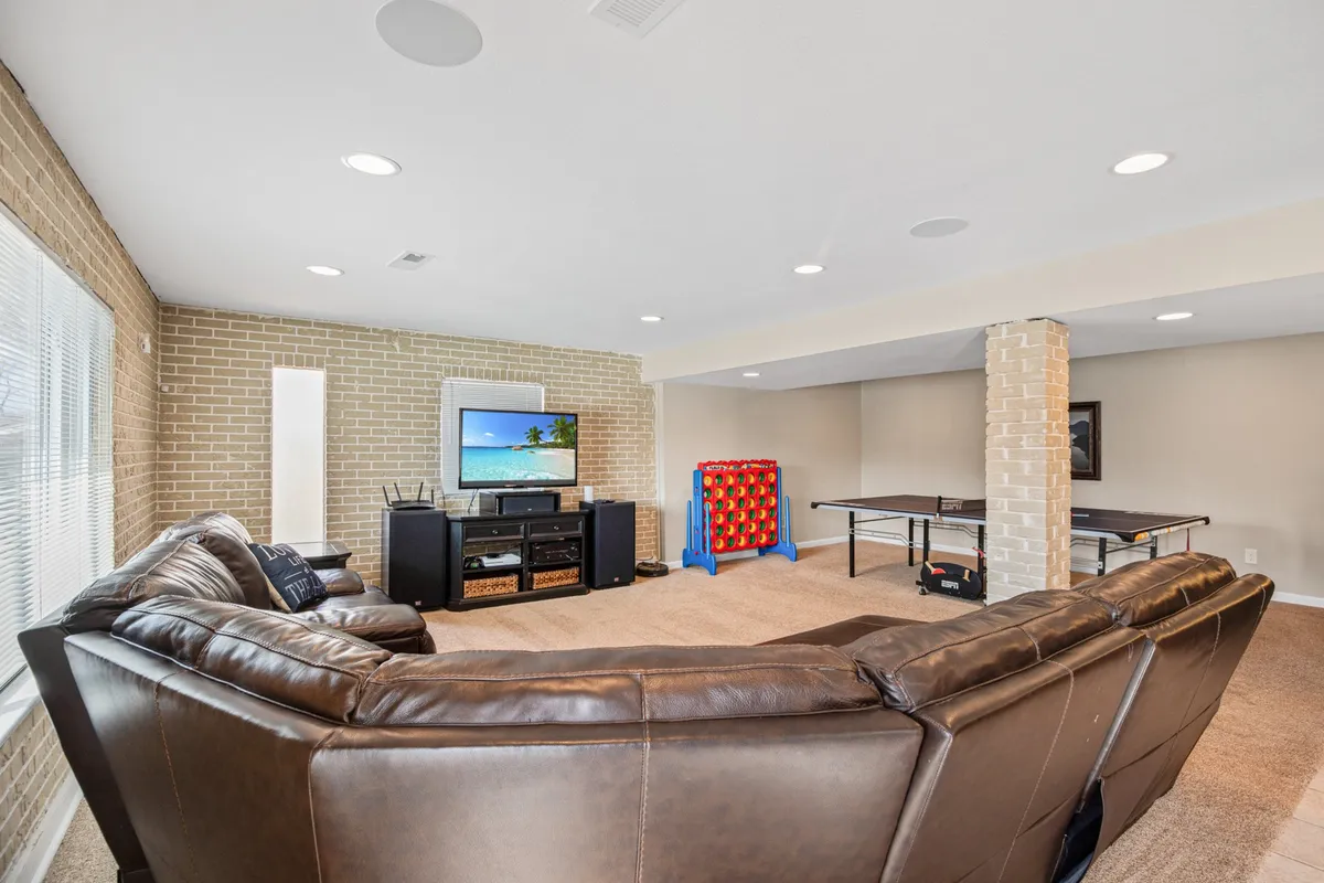 A vibrant, open living room featuring a large sectional sofa, a television displaying a scenic beach image, a ping pong table, and a colorful game table. The space is designed for leisure and entertainment, with plenty of seating and activity options for groups.