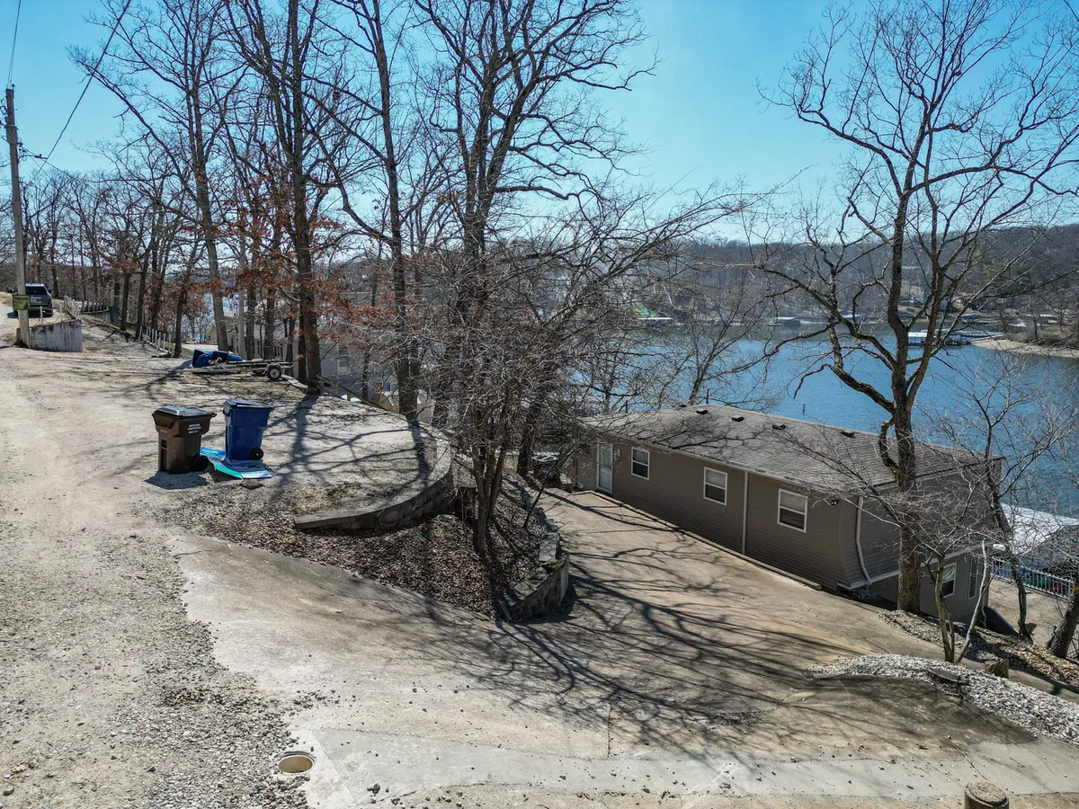 This exterior image captures a peaceful lakeside setting with bare trees lining a gravel path, the lake stretching into the distance, and a glimpse of the house nearby. The natural environment and tranquil water offer a clear sense of access to the outdoors.