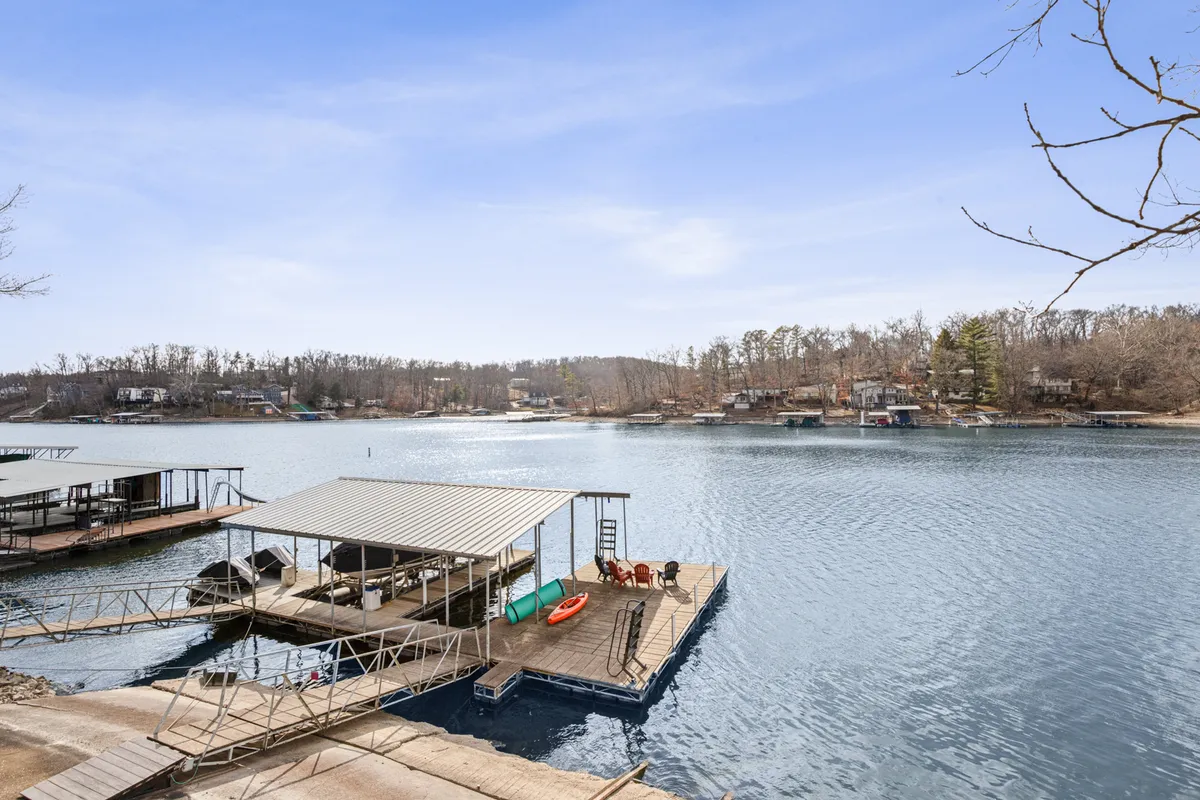 The dock is featured prominently, with a kayak resting nearby, water shimmering in the background, and houses dotting the shoreline. The setting suggests both active water recreation and tranquil relaxation.