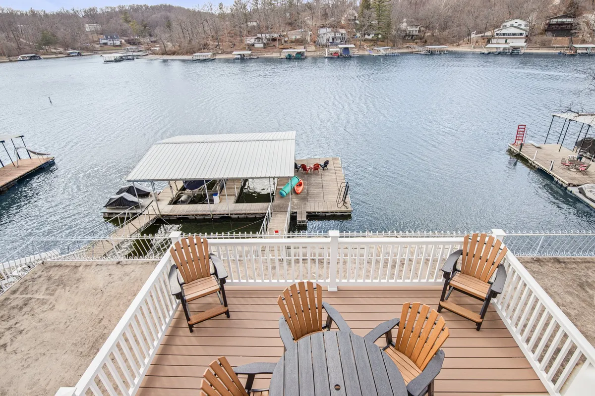 A picturesque dock extends over the calm lake, surrounded by lush greenery. Outdoor furniture is set up on the dock, creating a welcoming space for relaxing or fishing. The tranquil water and inviting dock are central in the composition, highlighting both the private and functional aspects of the space.
