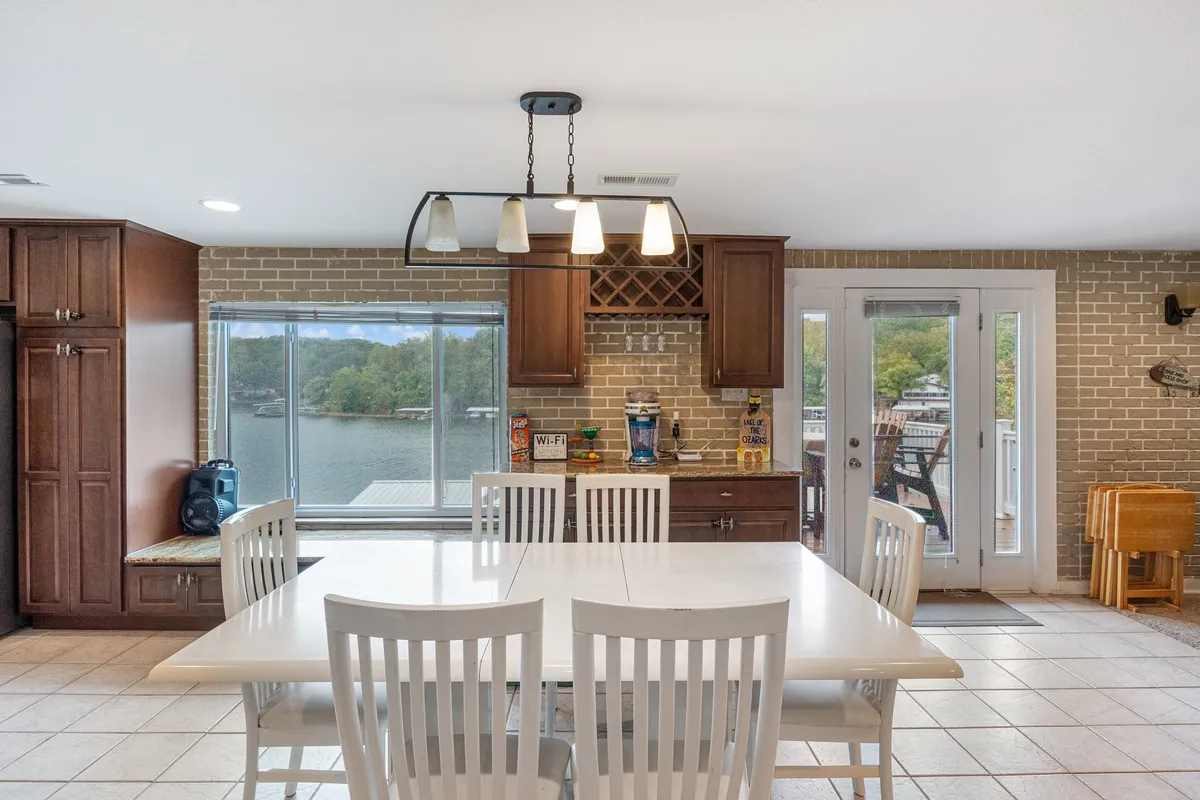 A warm dining area with a large wooden table positioned to enjoy gorgeous views of the lake through expansive windows. The room includes wooden cabinetry and visible kitchen appliances, creating a seamless space for cooking and dining together.