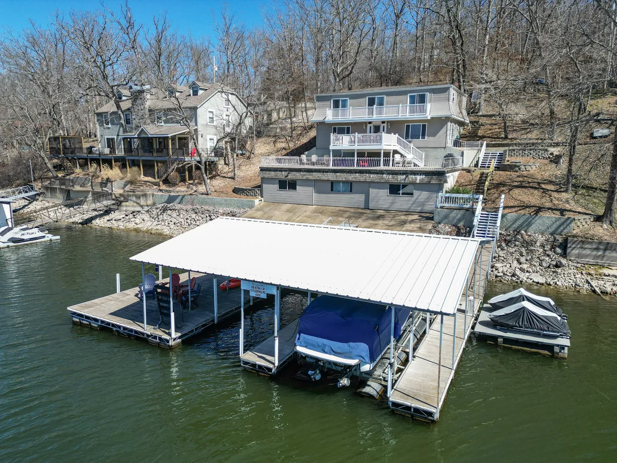 This image captures the private dock extending into a tranquil lake, with the house visible on a gentle hill and boats docked nearby. The setting is peaceful, surrounded by lush greenery, evoking a sense of seclusion and family-friendly adventure.