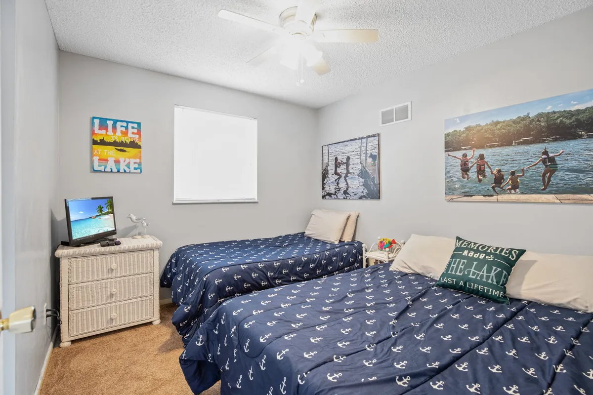 A cheerful bedroom featuring two twin beds with anchor-patterned bedding, a wicker nightstand between them, and wall art with lake scenes and motivational quotes. The space feels welcoming and is ideal for children or multiple guests.
