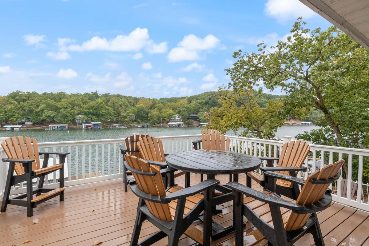 A welcoming outdoor dining set is positioned on a spacious deck overlooking the serene lake and surrounded by trees, suggesting a perfect spot for family meals, games, and relaxation with beautiful water views.