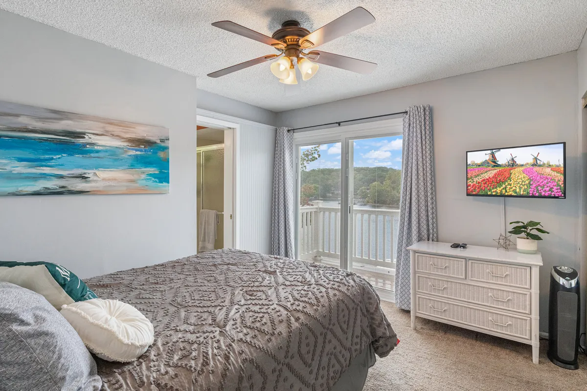 A serene bedroom with a plush bed covered in textured bedding, a large window offering peaceful views, artwork hanging on the wall, a TV set on the dresser, and a leafy potted plant adding a touch of freshness. The atmosphere is tranquil and inviting, perfect for relaxation.