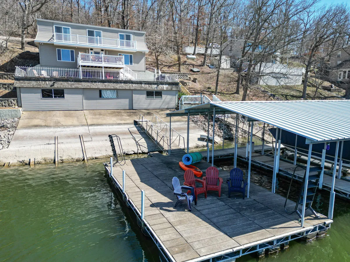 A wide shot shows the house with a balcony, a brightly colored dock with chairs, and calm lake water. The dock looks lively and well-equipped, perfect for family gatherings and relaxing at the water’s edge.