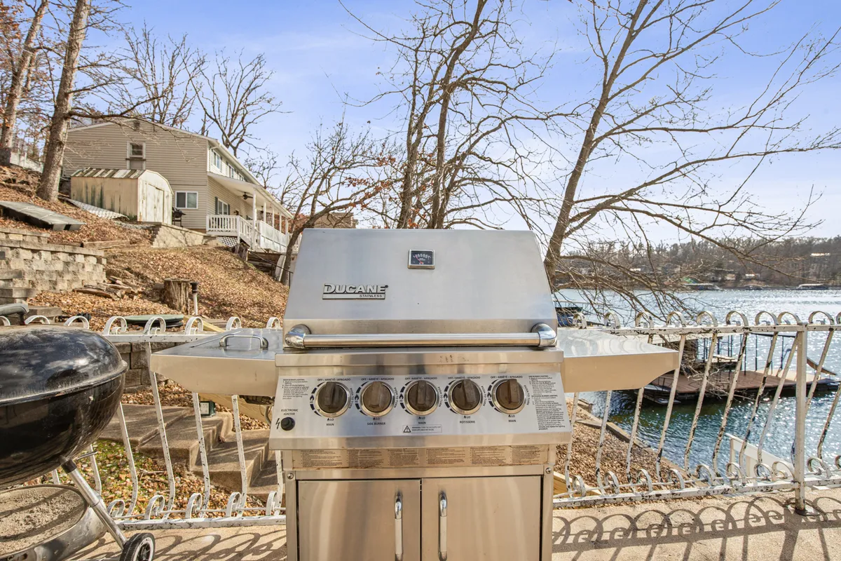 This exterior scene spotlights a grill situated on a deck by the waterfront, with views of the lake, distant houses, and leafy trees. The setup hints at opportunities for barbecues, outdoor dining, and lively gatherings in a scenic setting.